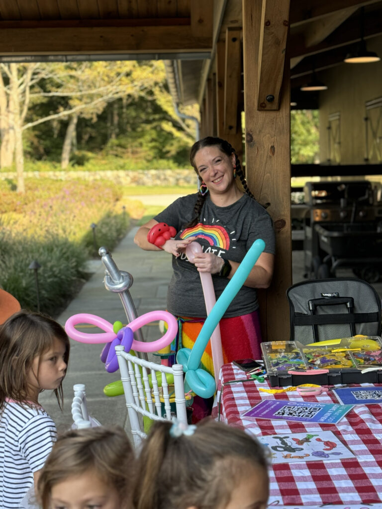 A balloon twister smiling at the camera while she twists a flower. The event is outdoors and there are kids in line.