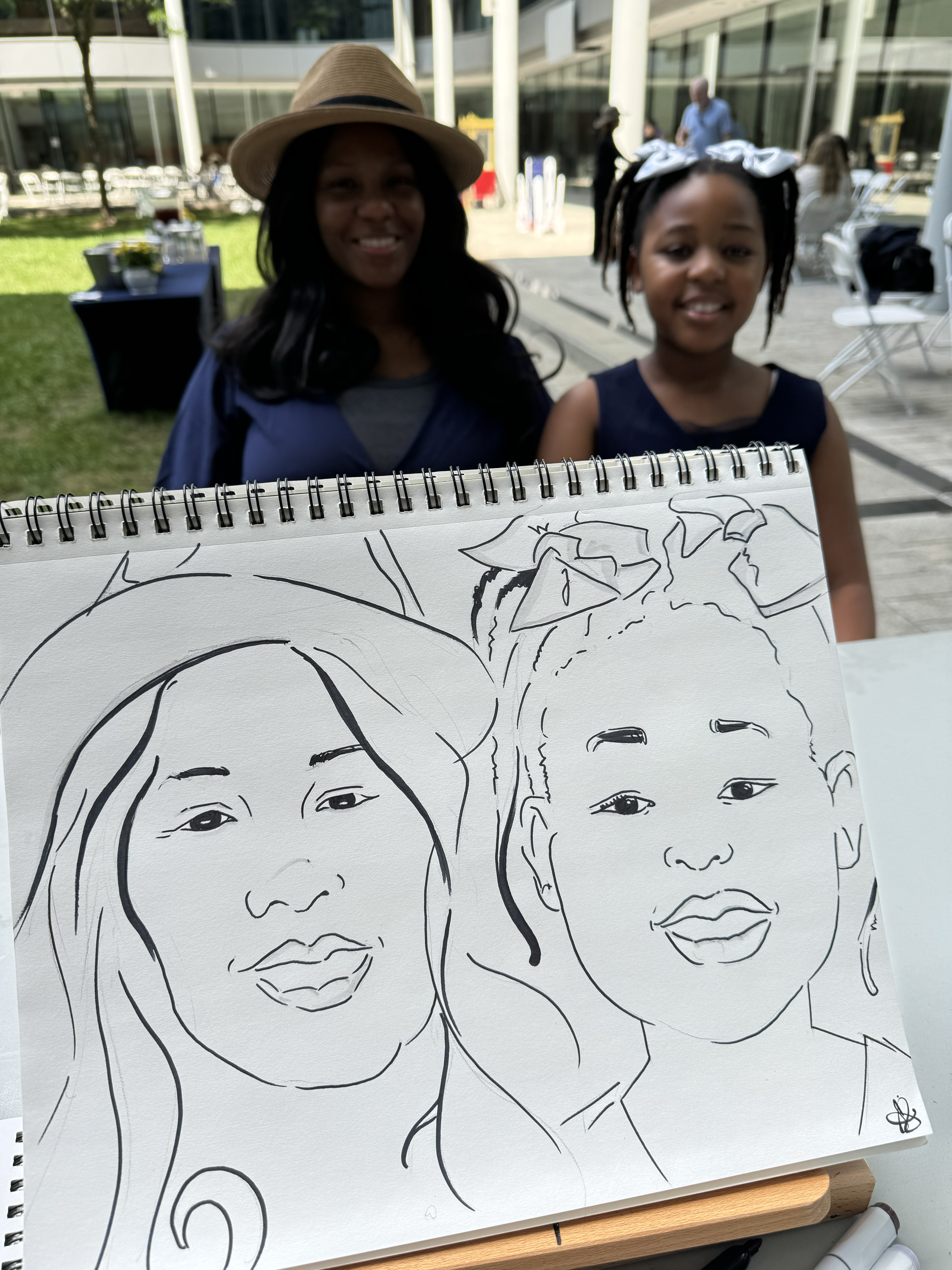 A mother and daughter sitting to have their caricatures drawn in RI. The POV is by the caricature artist, and you can see both the family and the drawing in the photo.