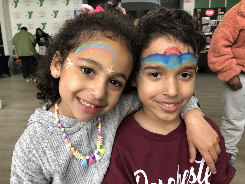 Two children, one with a rainbow and one with a batman design, face painted by The Face Painter RI