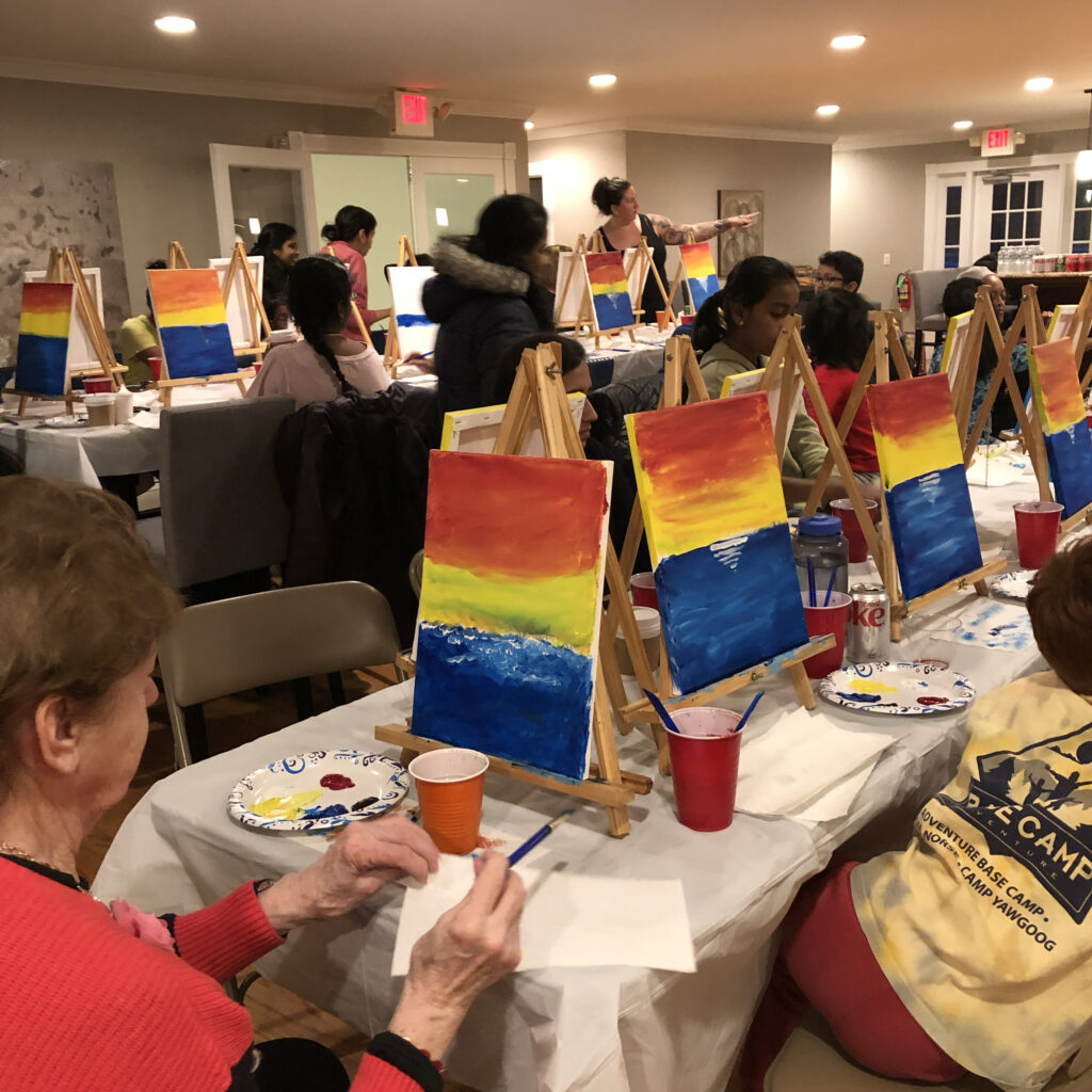 A large group at an indoor event doing a paint night together. The teacher from Face Painter RI is in the background pointing to an example, which the adults are looking at.