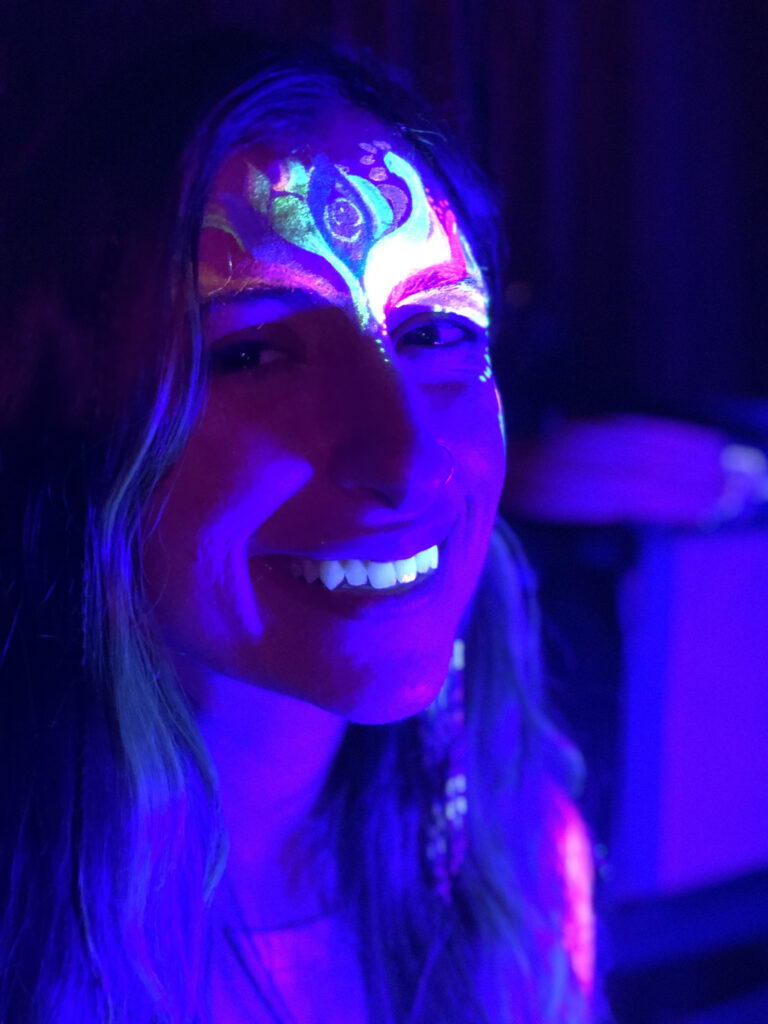 A woman laughing at the camera indoors in a dark room with a glowing face paint design