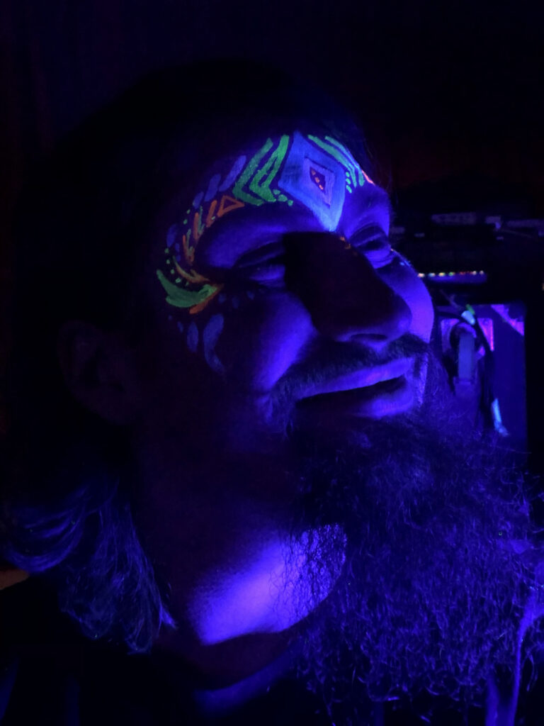 A man with a beard smiling with a glow in the dark face paint design. The room is dark and the face painting is glowing.